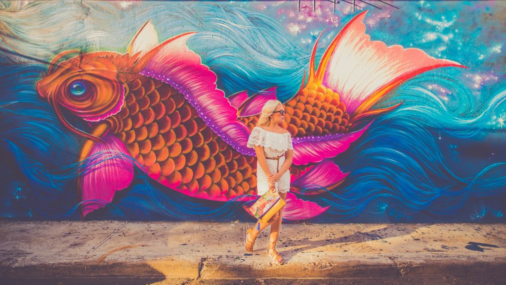 Une femme en vêtements d'été blancs et lunettes de soleil se tient devant une fresque murale colorée représentant un grand poisson koi vibrant